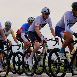 A group of professional road cyclists riding in a peloton during a race, wearing white, blue, and yellow team jerseys, helmets, and sunglasses on drop-barred racing bicycles.